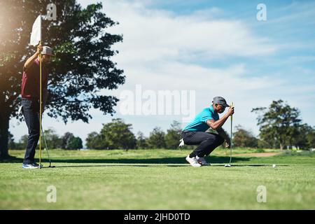 Dieses wird innen sein. Ein fokussierter junger männlicher Golfspieler, der einen Golfball betrachtet, während er auf dem Gras draußen während des Tages sitzt. Stockfoto