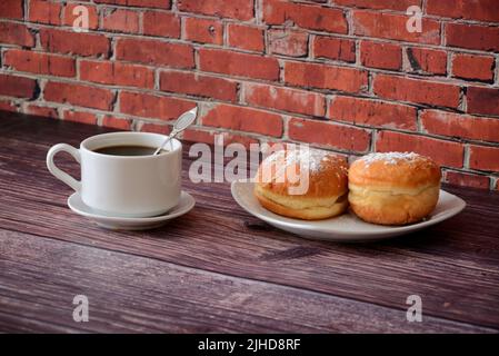 Eine Tasse heißen Kaffee auf einer Untertasse mit einem Löffel und einem Teller mit zwei Donuts, die mit Puderzucker bedeckt sind, auf einem Holztisch. Nahaufnahme. Stockfoto