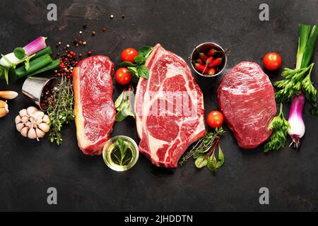 Verschiedene Steaks aus rohem Rindfleisch zum Grillieren mit Gewürzen und Geschirr auf dunklem Hintergrund, Draufsicht. Stockfoto