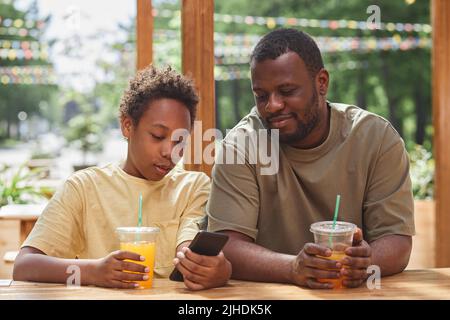 Afrikanischer Junge, der seinem Vater Videos oder Fotos auf seinem Smartphone zeigt, während sie im Café im Freien Saft trinken Stockfoto