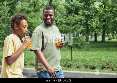 Glücklicher afrikanischer Vater, der während der Sommerferien kalten Saft trinkt und mit seinem Sohn im Park spazieren geht Stockfoto