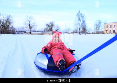 Ein Mädchen in einem Winteroverall sitzt auf einem Schlauch. Das Kind ist im Schnee Schlittenfahren. Stockfoto