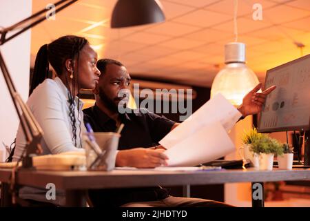 afroamerikanischer Mann und Frau, die im Team zusammenarbeiten, um einen Präsentationsbericht zu erstellen und Daten auf Computern und Papieren zu betrachten. Zusammenarbeit mit Papierkram im Büro während des Sonnenuntergangs. Stockfoto
