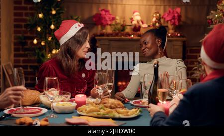 Eine aufgeregte Frau, die beim Weihnachtsessen mit einem engen Freund diskutiert, während sie Champagner trinkt. Festliche, vielfältige Menschen feiern das Winterfest mit Sekt und traditionellen hausgemachten Speisen. Stockfoto