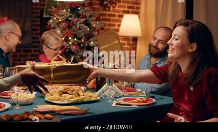 Festliche Familienmitglieder sitzen am Weihnachtstisch, während sie sich zu Hause mit Geschenken austauschen. Fröhliche Menschen versammelten sich im Wohnzimmer, um den traditionellen Winterurlaub zu feiern, während sie einander Geschenke gaben. Stockfoto