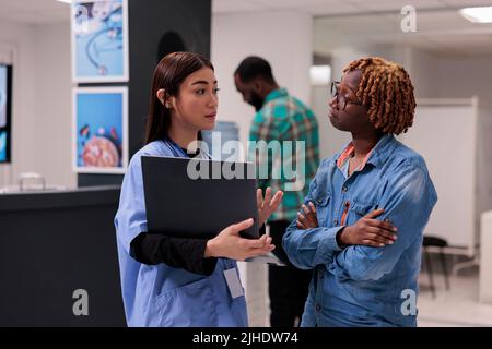 afroamerikanische Frau und Krankenschwester, die in der Lobby der Einrichtung auf einen Laptop schauen, um eine Behandlung zur Gesundheitsdiagnose zu finden. Menschen, die über Krankheit und Medizin sprechen, benutzen Computer an der Rezeption. Stockfoto