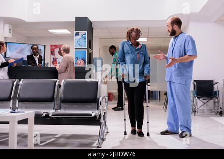 Junge Frau mit Krücken, die in der Lobby der medizinischen Einrichtung mit einer Krankenschwester spricht und auf eine Gesundheitsberatung mit einem Spezialisten wartet. Frau, die an der Krankenhausaufnahme mit körperlichen Beeinträchtigungen zu tun hat. Stockfoto