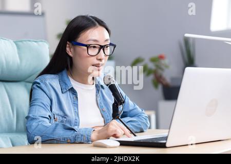 Podcast-Aufzeichnung, Interview. Junge schöne asiatische Frau, die mit einem Mikrofon und einem Laptop an einem Tisch sitzt. Spricht, erzählt. Macht eine Audioaufnahme eines Podcasts, gibt ein Interview per Videoanruf. Stockfoto