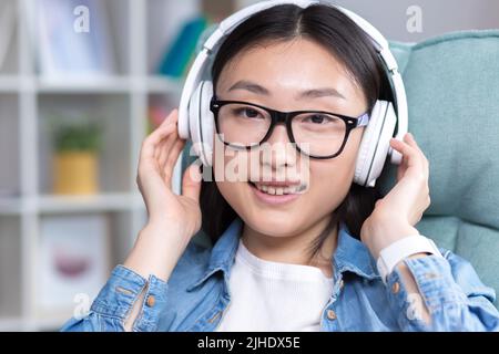 Die junge, schöne asiatische Frau, die mit Kopfhörern Musik hört, die Augen schließt und ihre Hände hinter ihren Kopf wirft, ruht, blickt auf die Kamera und grinst Stockfoto