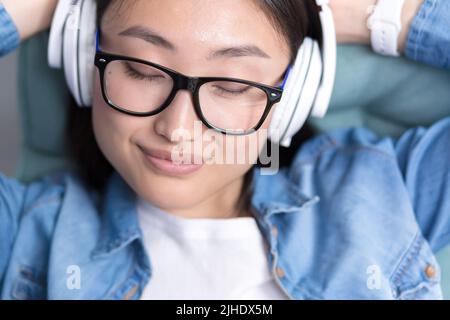 Die junge, schöne asiatische Frau, die in Kopfhörern Musik hört, die Augen schließt und ihre Hände hinter den Kopf wirft, ruht Stockfoto