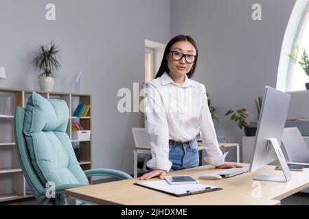 Porträt einer jungen schönen asiatischen Frau in Brille Geschäftsfrau, Manager, Regisseur, Chef im Büro am Schreibtisch stehen. Sie lehnt sich mit den Händen auf den Tisch, blickt lächelnd auf die Kamera. Stockfoto