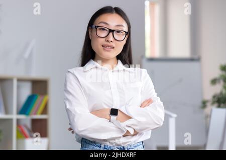 Nahaufnahme. Porträt einer jungen schönen asiatischen Frau in Brille Geschäftsfrau, Manager, Regisseur, Chef im Büro am Schreibtisch stehen. Kreuzte ihre Arme, schaut lächelnd auf die Kamera. Stockfoto