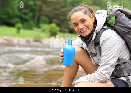 Ein glücklicher Wanderer, der eine Kantine hält, sieht Sie an einem Flussufer im Berg an Stockfoto