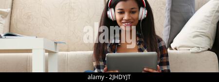 Lateinische junge Frau sitzt mit Tablet auf dem Teppich und schaut sich die Show an, Dame im Headset Stockfoto