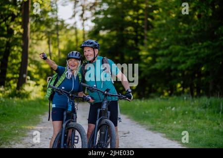 Porträt eines aktiven älteren Paares, das im Sommerpark Fahrrad fährt, auf dem Weg steht und die Kamera anschaut Stockfoto