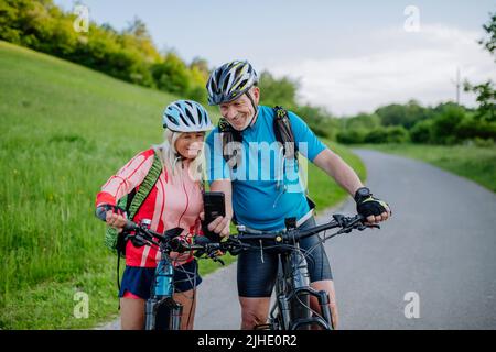 Aktives Seniorenpaar, das sich nach einer Fahrradtour im Sommerpark mit dem Smartphone ausruhte. Stockfoto