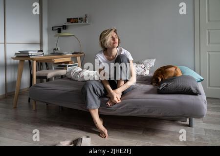 Einsamkeit in der Mitte des Lebens. Nachdenkliche, traurige Frau mittleren Alters, die auf dem Sofa sitzt und einen Hund in der Ferne sieht Stockfoto