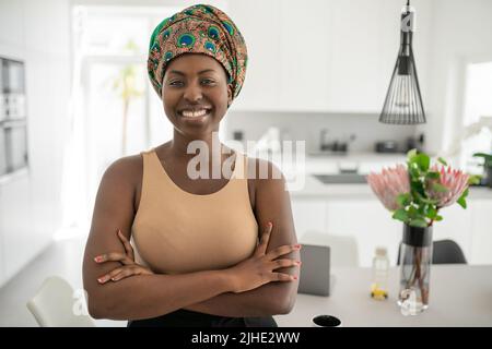 Porträt einer selbstbewussten, lächelnden Afrikanerin mit gekreuzten Armen und Blick in die Kamera. Arbeiten von zu Hause aus und tragen ein traditionelles Kopftuch Stockfoto
