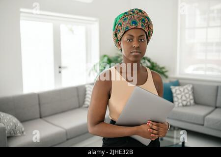 Porträt einer schönen, lächelnden schwarzafrikanischen Frau, die in einem modernen Zuhause mit einem Laptop in der Hand steht und ein traditionelles Kopftuch trägt Stockfoto