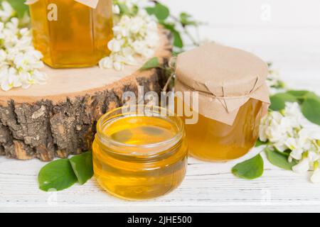 Süßes Honigglas umgab die Akazienblüten des Frühlings. Honig fließt aus einem Löffel in ein Glas. Gläser von klarem frischen Akazienhonig auf Holzgrund. Stockfoto