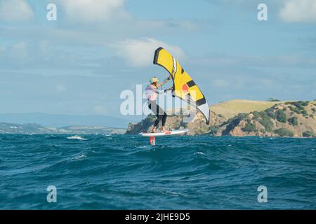 Ein Mann Flügel Folien auf dem Meer mit einem Hand gehalten aufblasbaren Flügel, Reiten ein Tragflächenboot Surfboard Flügel Folie Bord. Hauraki Gulf, Auckland, Neuseeland. Stockfoto