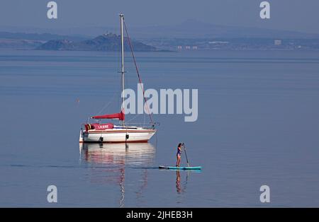 Portobello, Edinburgh, Schottland, Großbritannien. 18.. Juli 2022. Die Temperatur stieg mit 23 Grad Celsius nur after10am, stieg bis 1pm sogar auf 29 Grad Celsius. Im Bild: Cooler Paddlebarder, der den ruhigen Firth of Forth mit Inchkeith Island im Hintergrund genießt. Stockfoto