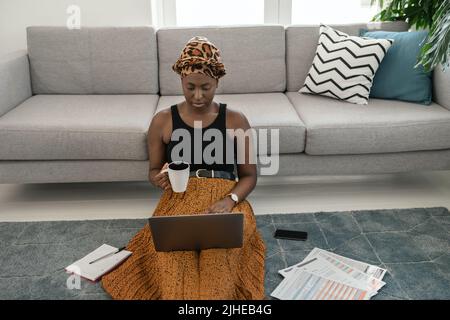 Schöne schwarze afrikanische Frau, die in der Lounge auf dem Boden sitzt und zu Hause mit einem traditionellen Kopftuch auf einem Laptop studiert Stockfoto