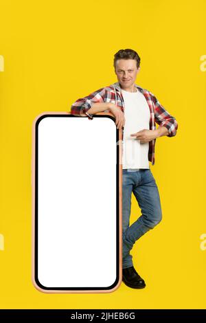 Hübscher junger Mann, der auf einem riesigen, riesigen Smartphone, einem digitalen Tablet mit weißem, schwarzem Bildschirm, der auf ein kariertes Hemd und Jeans auf gelbem Hintergrund zeigt, steht. Modell mit freiem Speicherplatz. Stockfoto