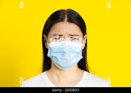 Wütend, traurig über Pandemie reife asiatische Frau trägt medizinische Gesichtsmaske Coronavirus oder Monkeypox Prävention. Charmante Frau mittleren Alters in weißem T-Shirt und medizinische Maske auf gelbem Hintergrund. Stockfoto