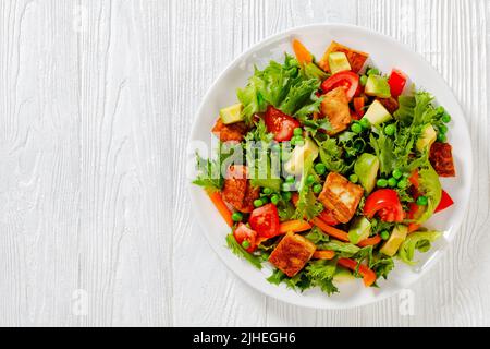 Tofu-Salat mit Avocado, knackigen Salatblättern, grünen Erbsen, Tomaten, gelber Paprika auf weißem Teller auf weißem Tisch, horizontale Ansicht von oben, Stockfoto