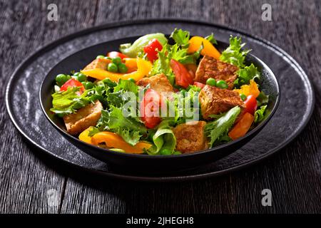 Gebratener Tofu-Salat mit knackigen Salatblättern, grünen Erbsen, Tomaten, gelbem Paprika in schwarzer Schüssel Stockfoto