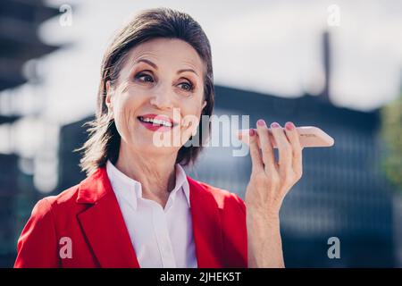Foto von positiven aufmerksam reifen Dame tragen rote Jacke hören Sprachnachricht modernes Gerät außerhalb der Stadt Business-Komplex Stockfoto