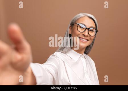 Die Dame, die eine Brille trägt, lächelt und schaut isoliert auf die beige Kamera. Fröhliche ältere asiatische Frau, die Selfie nimmt. POV Stockfoto