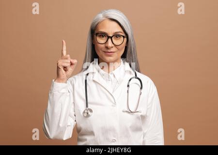 Lächelnde reife grauhaarige asiatische Ärztin in weißem medizinischen Kleid, mit Stethoskop isoliert auf braunem Hintergrund zeigt den Finger nach oben. MD achtet auf Krankenversicherung und Krankenversicherung Stockfoto