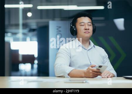 Der asiatische Chef-Unternehmer entspannt sich im Büro, hört entspannende und entspannende Musik, Mann sitzt am Schreibtisch, mit großen Kopfhörern und Online-Höranwendungen am Telefon Stockfoto