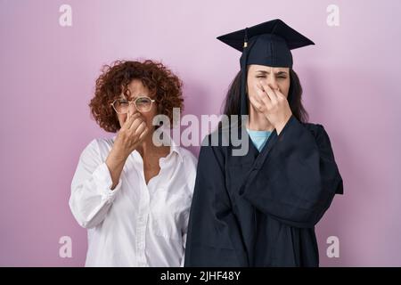 Hispanische Mutter und Tochter tragen Abschlusskappe und Zeremoniengewand und riechen etwas stinkendes und ekelhaftes, unerträglicher Geruch, halten Atem an Stockfoto