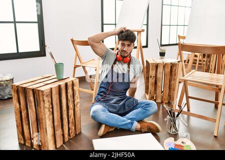 Junger hispanischer Mann, der im Kunststudio sitzt, verwirrt und wundert sich über Fragen. Unsicher mit Zweifel, mit der Hand auf dem Kopf denkend. Nachdenkliches Konzept. Stockfoto