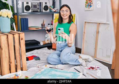 Junge hispanische Künstlerin, die im Kunststudio mit einem Smartphone Wein trinkt Stockfoto