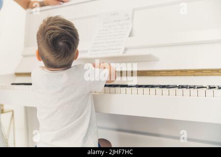 Kleiner nicht erkennbarer kaukasischer Junge in einem weißen T-Shirt, der vor einem wunderschönen elfenbeinweißen Klavier sitzt. Musik als Hobby für kleine Kinder. Hochwertige Fotos Stockfoto