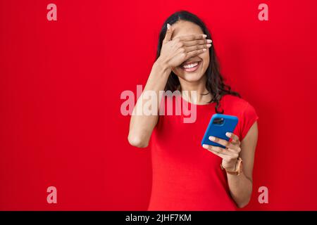 Junge brasilianische Frau mit Smartphone über rotem Hintergrund lächelnd und lachend mit Hand auf Gesicht bedeckende Augen für Überraschung. Blind Konzept. Stockfoto