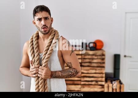 Junger hispanischer Mann, der im Schockgesicht mit Kampfseil im Fitnessstudio trainiert, skeptisch und sarkastisch aussackt und mit offenem Mund überrascht ist Stockfoto