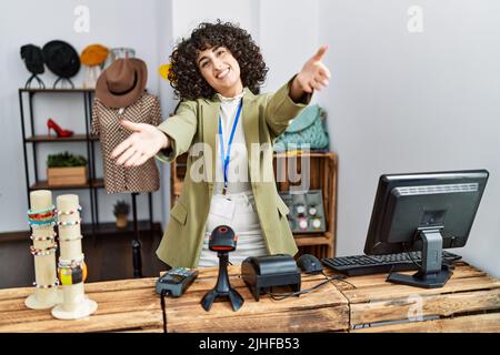 Junge Frau aus dem Nahen Osten, die als Managerin in einer Einzelhandelsboutique arbeitet und die Kamera lächelt und sich mit offenen Armen umarmt Stockfoto