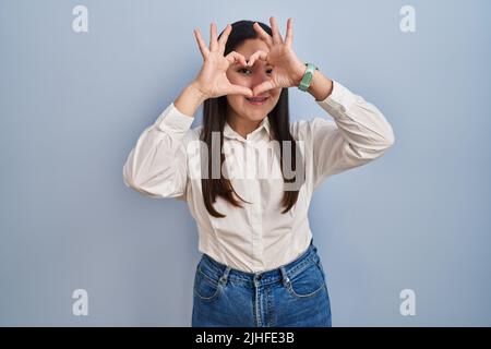 Junge lateinische Frau, die auf blauem Hintergrund steht und Herzform macht, wobei Hand und Finger lächelnd durch das Schild schauen Stockfoto