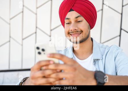 Fröhlicher indischer Kerl trägt traditionelle Turban Messaging, SMS auf dem Smartphone. Ein hinduistischer Student sitzt mit einer mobilen App im Freien, scrollt Nachrichten in den Feeds, websurft, spielt Spiele Stockfoto