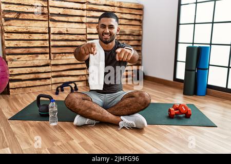 Junger inder, der auf der Trainingsmatte im Fitnessstudio sitzt und mit den Fingern auf Sie und die Kamera zeigt, lächelt positiv und fröhlich Stockfoto