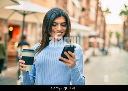 Junge hispanische Frau, die Smartphone benutzt und eine Tasse Kaffee in der Stadt trinkt Stockfoto
