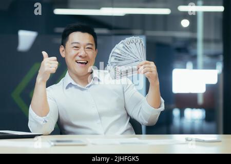 Porträt eines glücklichen asiatischen Geschäftsmannes im Büro mit Bargelddollar, lächelnder Mann und Blick auf die Kamera, der die Dollars in der Hand hält und den Daumen nach oben zeigt Stockfoto