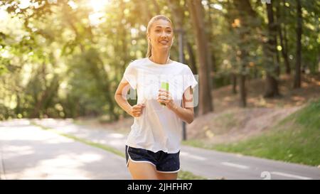 Frau hören Musik läuft auf Asphalt Spur Sommer Park kaukasischen Weibliche Kopfhörer Joggen sonnigen Morgen Läufer Training halten Smartphone in Handheiler Stockfoto