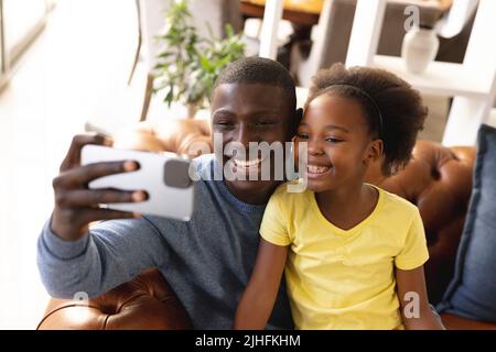 Bild eines glücklichen afroamerikanischen Vaters und einer Tochter, die auf dem Sofa sitzen und Selfie machen Stockfoto