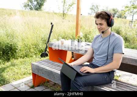 Cooler, junger Mann mit grauem Hemd sitzt draußen in einem Park mit Headset und arbeitet an seinem Tablet, im Hintergrund steht E-Scooter Stockfoto